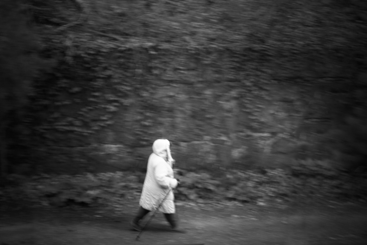 A solitary figure in a white coat walking briskly past a blurred hedge, suspended in a motion-filled monochrome landscape; a meditation on isolation and passage.