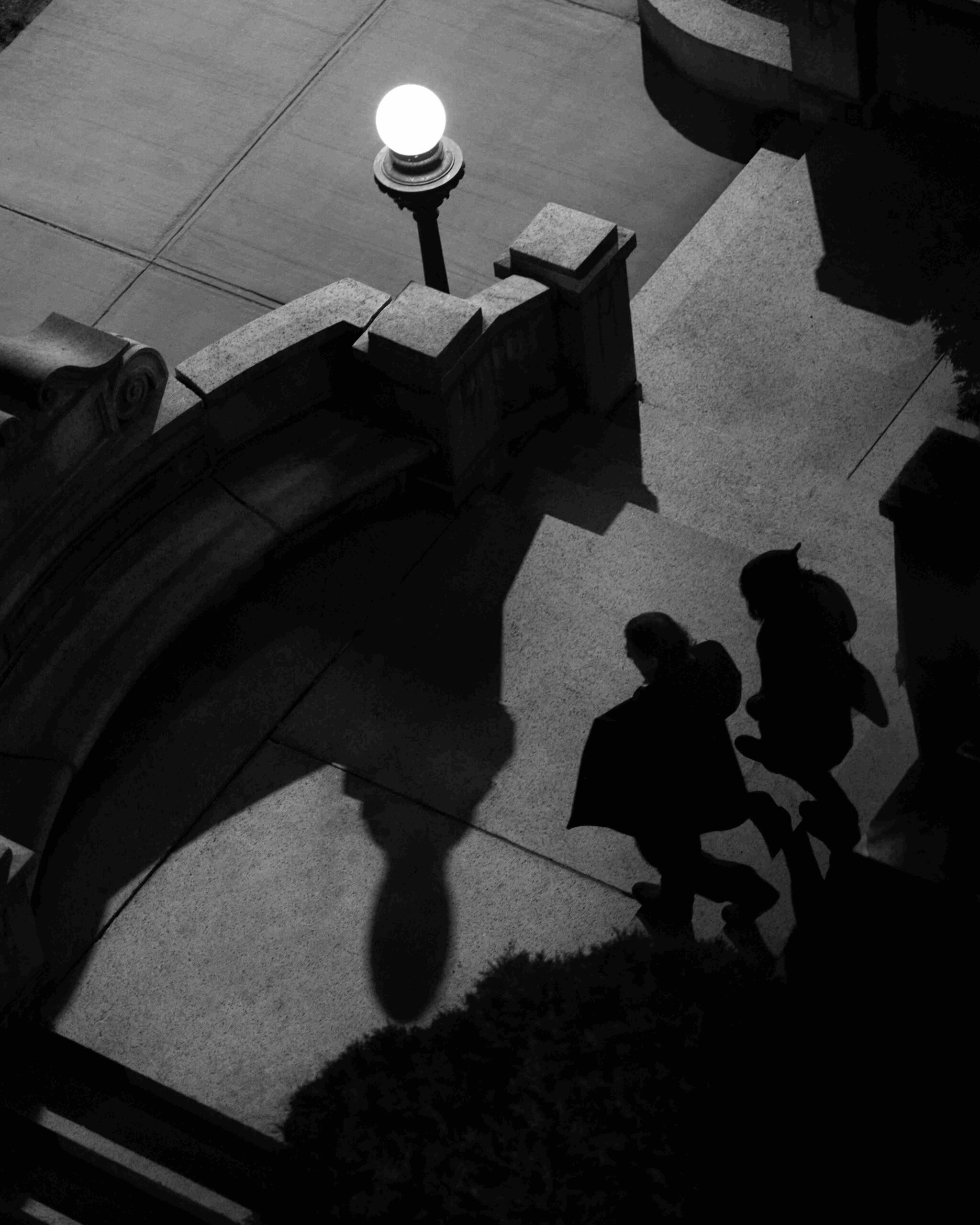 Overhead view of two silhouetted people ascending stone steps, elongated shadows cast in harsh light, suggesting private conversation and layers of presence.