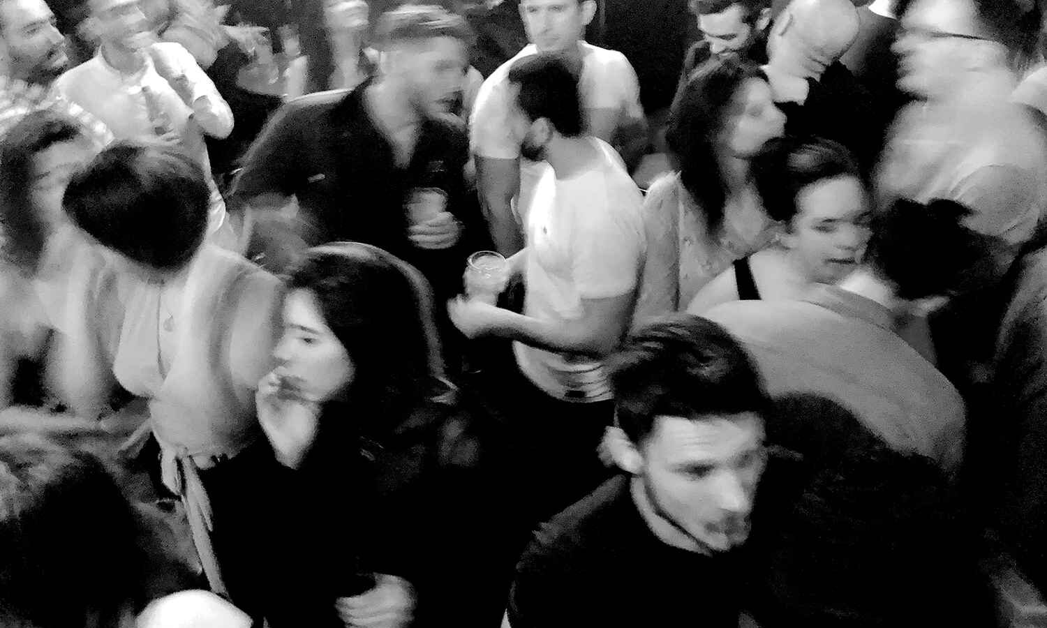 A dense, energetic crowd captured in black and white with faces turned, expressions and movements colliding to evoke the mystery of urban gatherings.