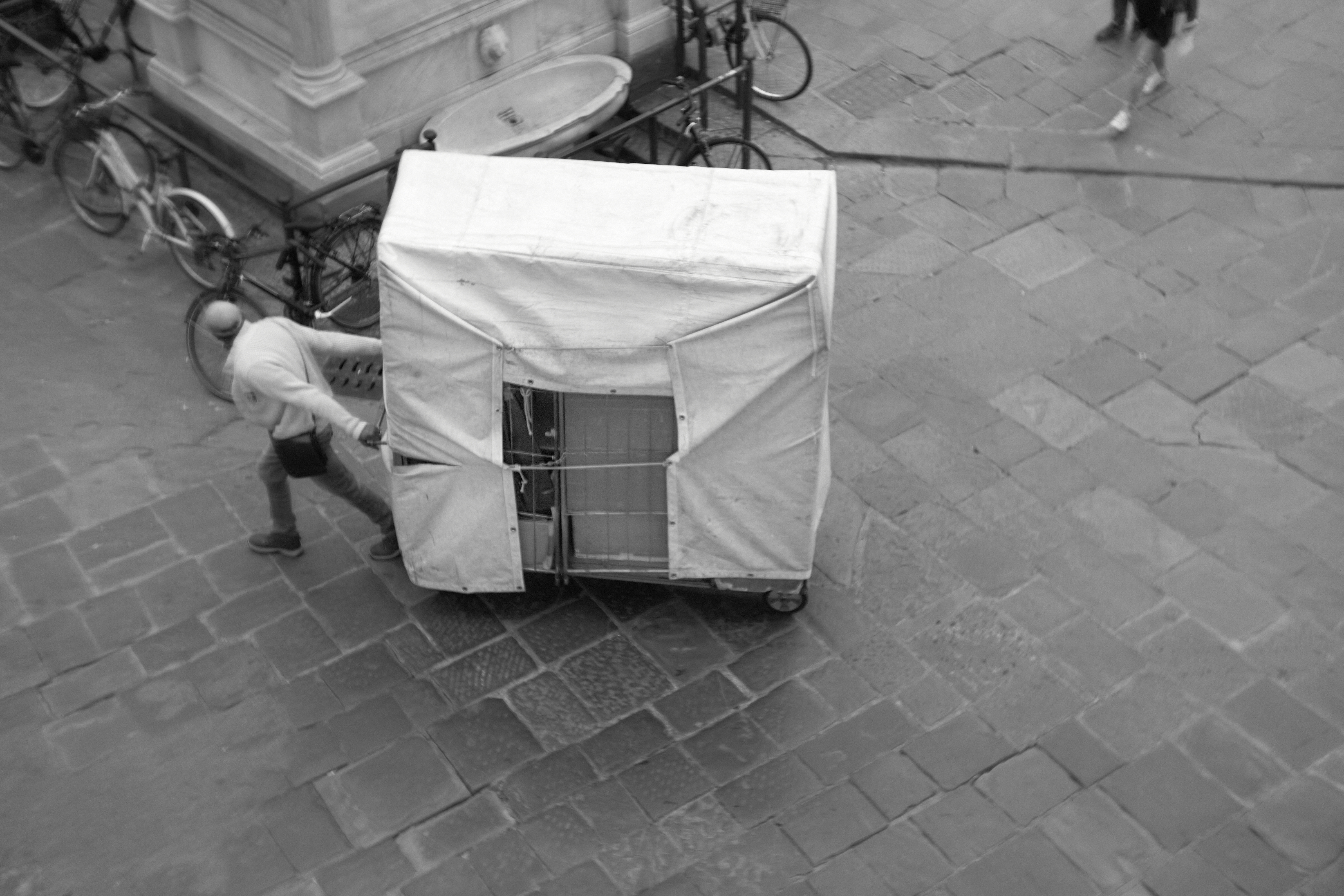 A figure bending beside a makeshift cart or shelter on pavement, the geometric structure and ambiguous intent conjuring themes of vulnerability and transient shelter.